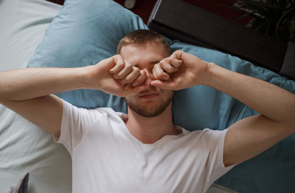 A young man lying in bed rubbing his eyes with his hands.
