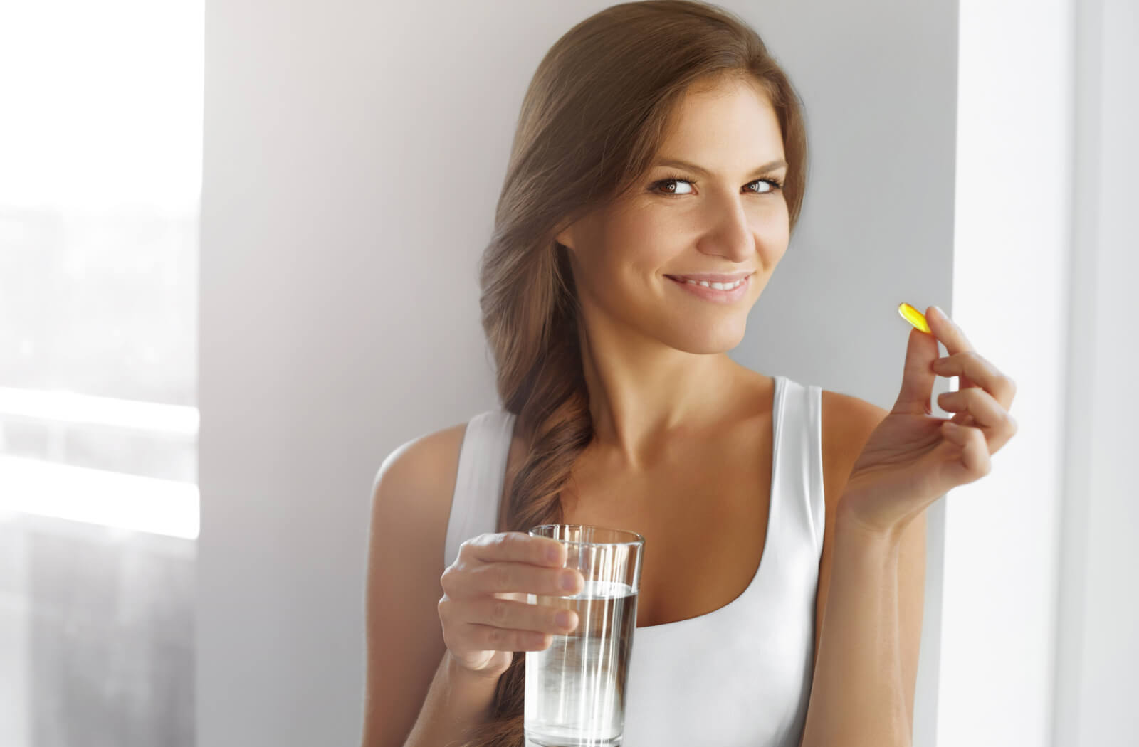 A smiling woman holding a vitamin pill in her left hand and a glass of water in her right hand