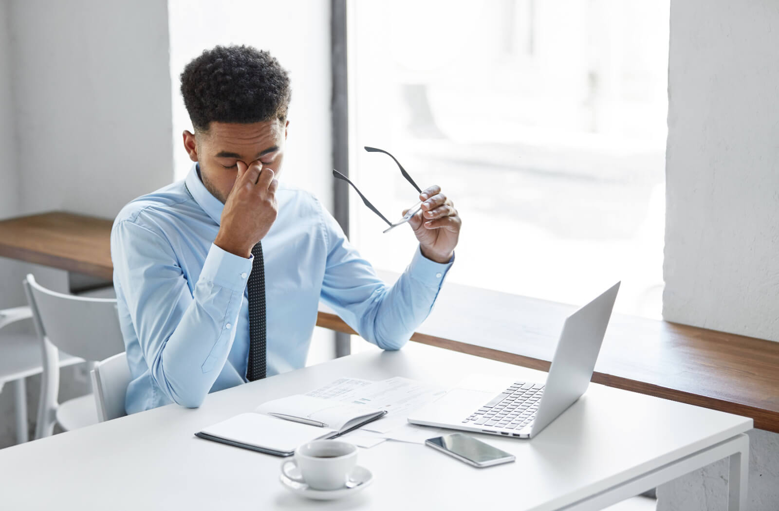 A young man sitting at a desk in an office with his laptop and holding his glasses in his left hand as he rubs his eyes