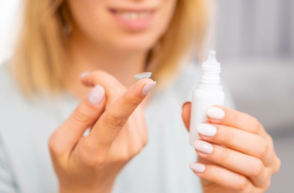 A close up view of a woman holding a contact lens on her right index finger. A bottle of eye drops is in her left hand.