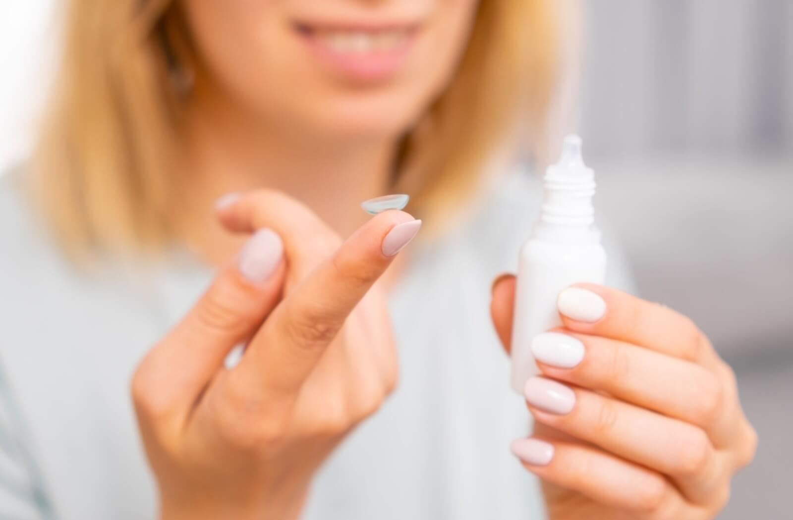A close up view of a woman holding a contact lens on her right index finger. A bottle of eye drops is in her left hand.