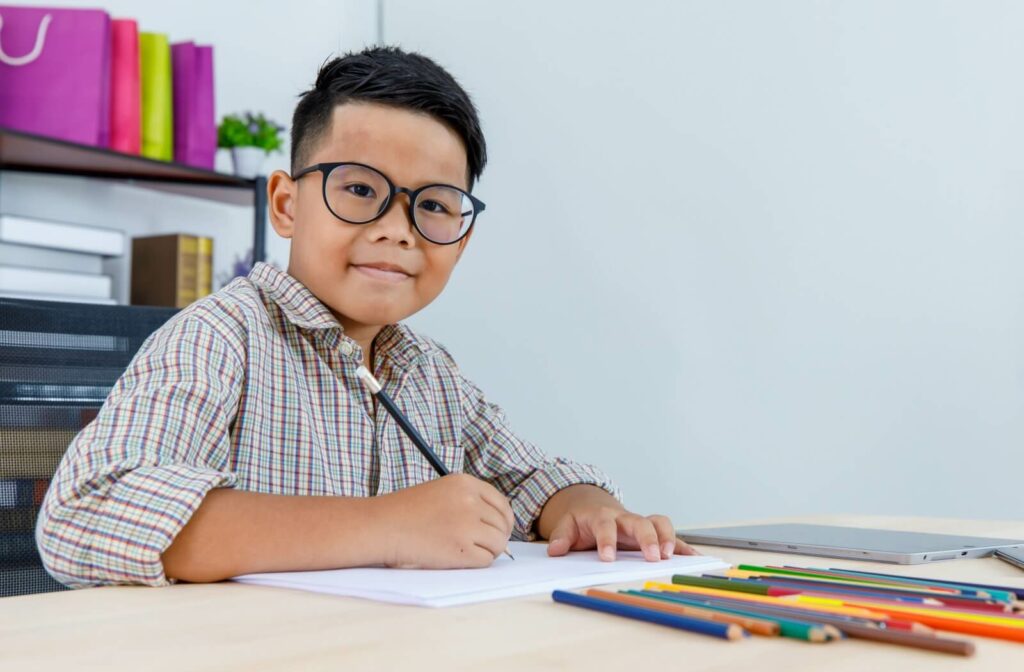 A young boy wearing glasses with myopia smiles as he does homework in a classroom.