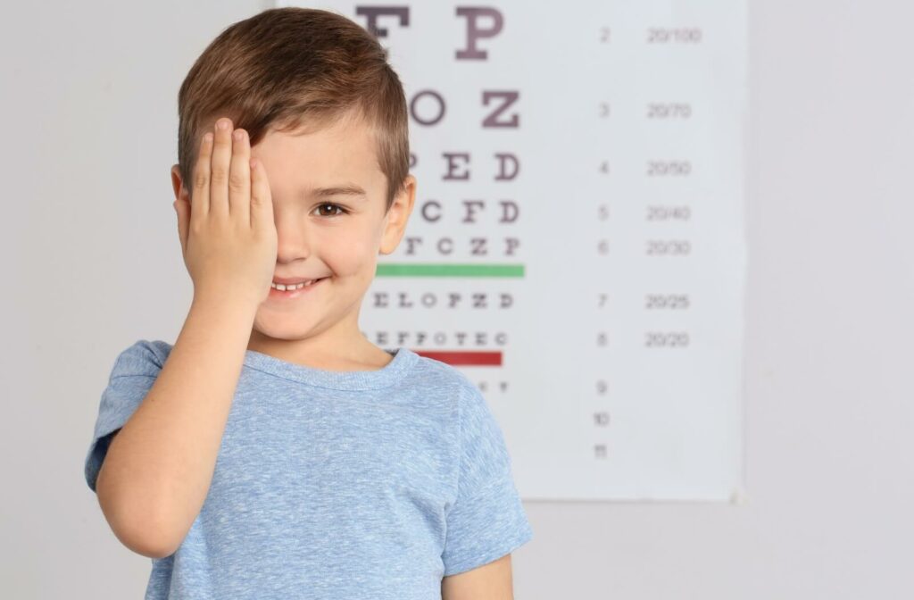 A young boy covering one eye stands in front of an eye chart and smiles.