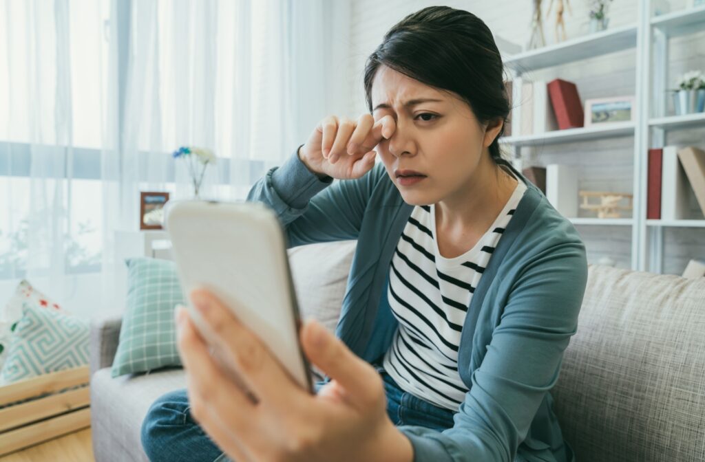 Person sitting on a couch, rubbing their eye while looking closely at a smartphone.