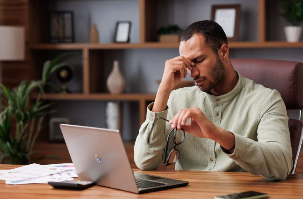 Person sitting at a desk holding their head and glasses, appearing fatigued while working on a laptop.