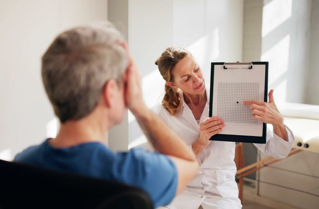 Optometrist in white lab coat holding up an Amsler grid, while an older adult covers one eye while looking at the chart.