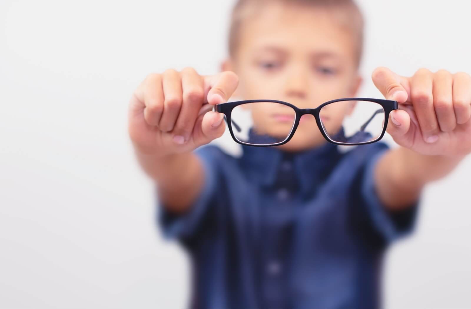 A young boy in a blue shirt holding a pair of black-rimmed glasses out in front of him, with the lenses in focus and his face blurred in the background.