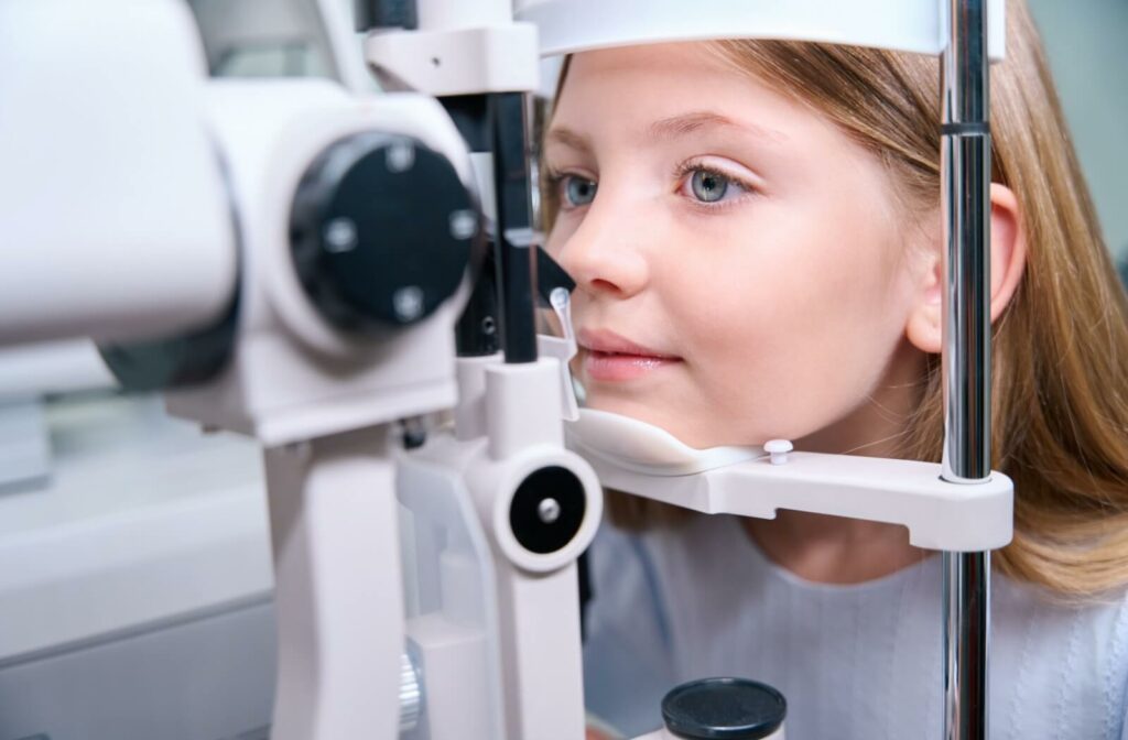 A young girl undergoing an eye exam, looking into a slit lamp machine at an optometry clinic, with her chin resting on the support bar.
