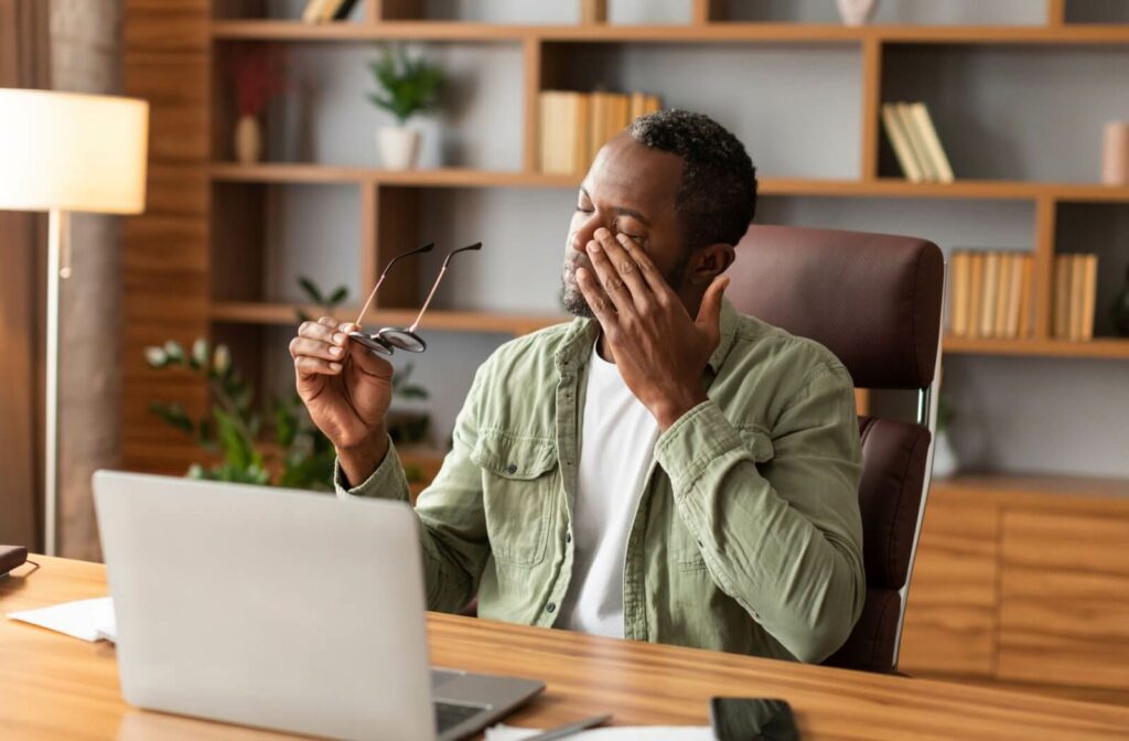 A person sitting at a desk removes their glasses and rubs their eyes in discomfort.