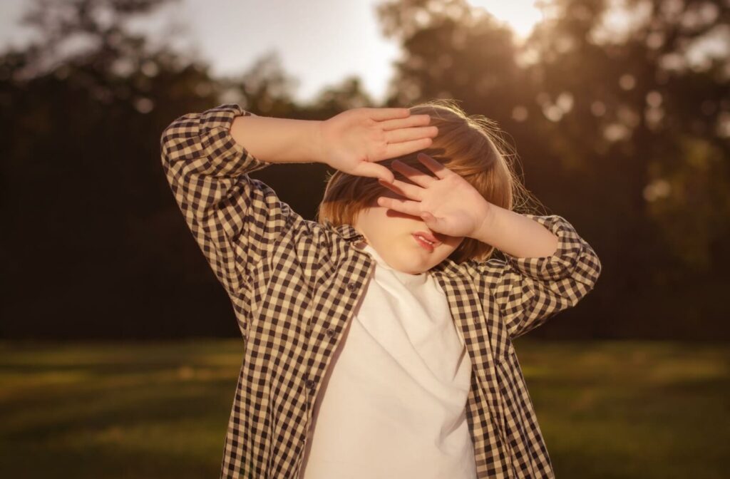 A child in a plaid shirt shielding their eyes with their hands against bright sunlight outdoors, illustrating photophobia or extreme light sensitivity.