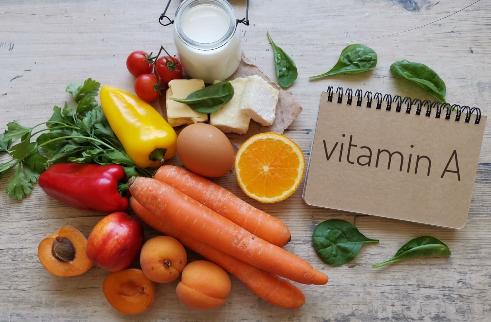 A flat lay of various vitamin A-rich foods, including carrots, bell peppers, eggs, and leafy greens, next to a notepad labeled "vitamin A" to highlight diet's role in eye health.