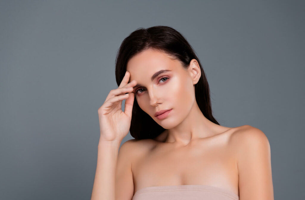 An individual with smooth, firm skin touching their forehead while posing against a gray background.